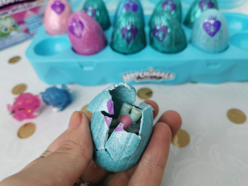 A hand holds a cracked open blue glittery egg, revealing a small toy creature inside. In the background, uncracked blue and purple eggs sit in a blue plastic tray, with two small toy creatures and gold confetti on a white surface nearby.