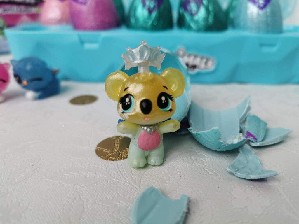 A small yellow and green toy bear with a pink belly and a silver crown stands on a white surface. Blue plastic eggshell pieces are broken nearby. In the background are other colourful toy animals and a blue container. Two gold coins lie on the table.