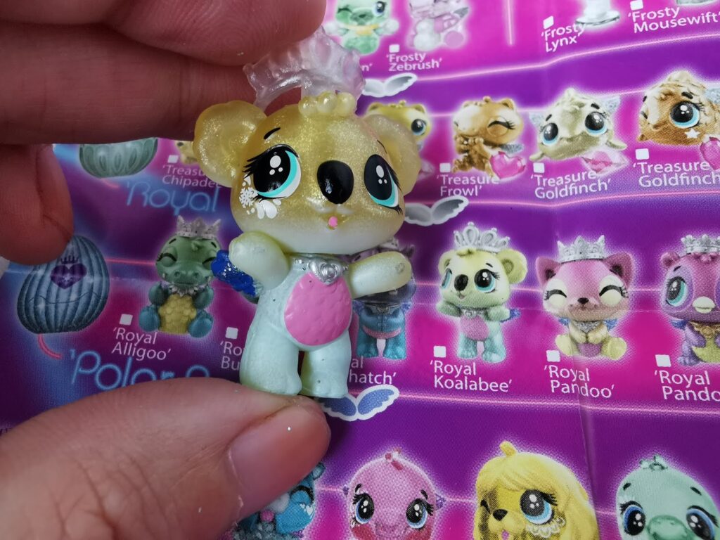 A hand holds a small toy koala with a crown and pink belly in front of a colourful collector’s guide featuring images and names of similar cute animal figurines on a purple background. The guide includes characters like “Royal Koalabee” and “Treasure Goldfinch”.