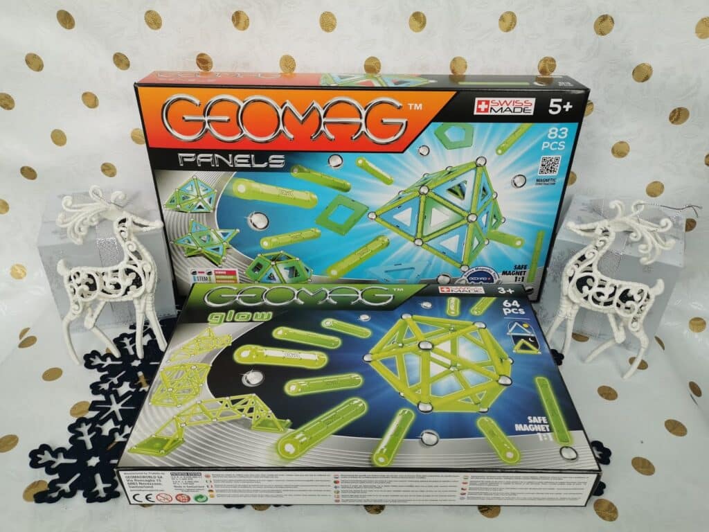Two boxed Geomag magnetic construction sets, one with 83 pieces and one with 64 pieces, are displayed upright and flat. The boxes feature green and yellow geometric shapes. Two decorative white reindeer and gold-patterned backdrop are in the background.
