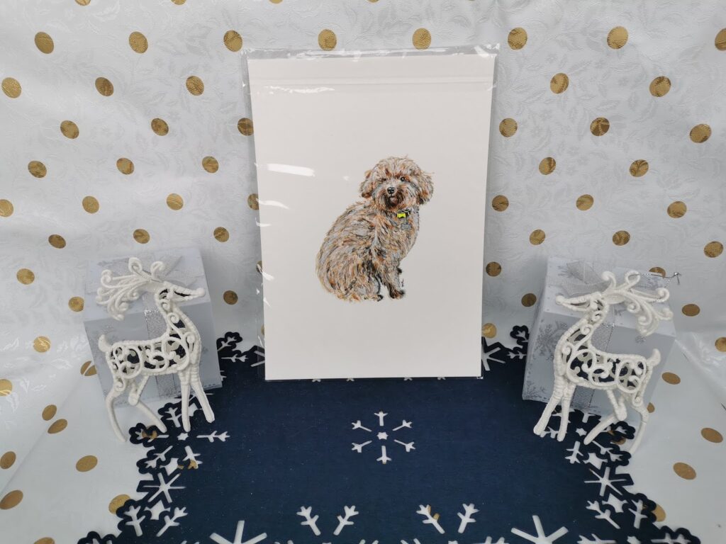 A framed drawing of a curly-haired dog sits upright on a dark blue mat with white snowflakes. The background is white with gold polka dots. Two white decorative reindeer with filigree designs stand on either side of the picture.