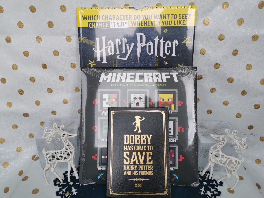 A Harry Potter-themed display with a yellow sign, a Minecraft 2020 calendar, and a black and gold notebook reading “Dobby has come to save Harry Potter and his friends 2020.” Two white reindeer decorations stand on either side, all on a star-patterned background.