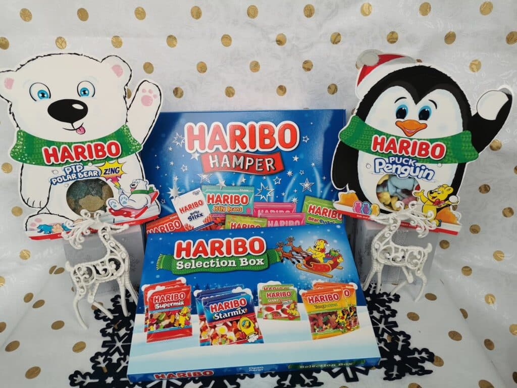 A festive Haribo display features a blue hamper box, a Haribo selection box, and various sweet packs, with large cut-outs of a polar bear and a penguin on either side, all set against a white and gold polka dot background with small white reindeer figures.