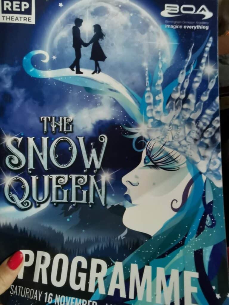 A theatre programme cover for "The Snow Queen" shows a stylised icy queen’s face in profile, adorned with a crystal crown. Silhouettes of a boy and girl stand on a moonlit bridge above her. The background is deep blue with snowflakes and sparkling stars.