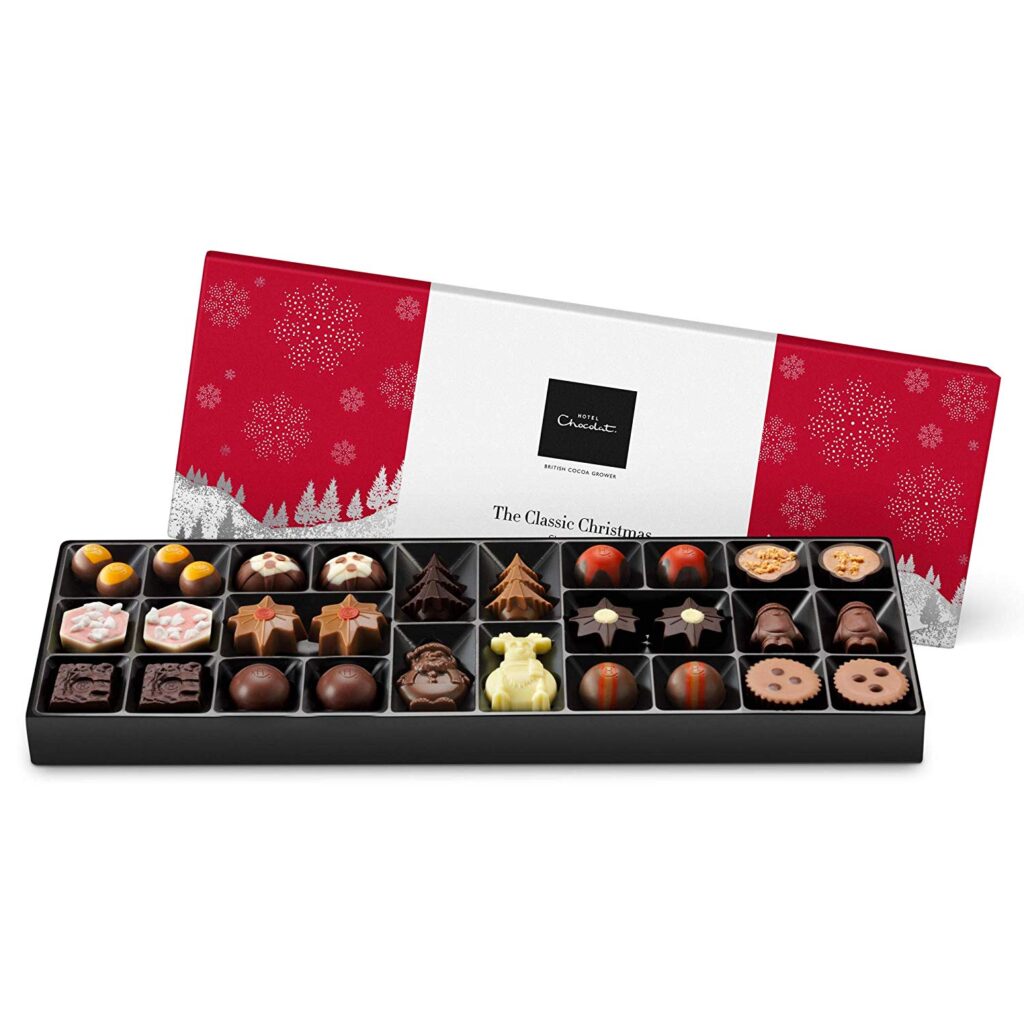 A rectangular box of assorted chocolates sits open, revealing 27 decorated chocolates in festive shapes like stars, Christmas trees, snowflakes, and round truffles. The box has a red and white holiday-themed lid with snowflake and tree designs.