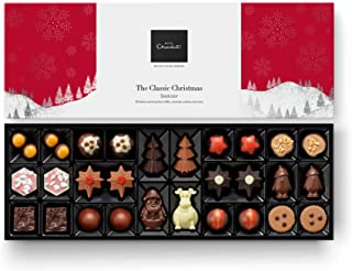 A festive chocolate box with assorted chocolates in holiday shapes—stars, trees, snowmen, Santa hats, and round truffles—arranged in a black tray. The box has a white background with red, snowflake-decorated panels and a winter scene.