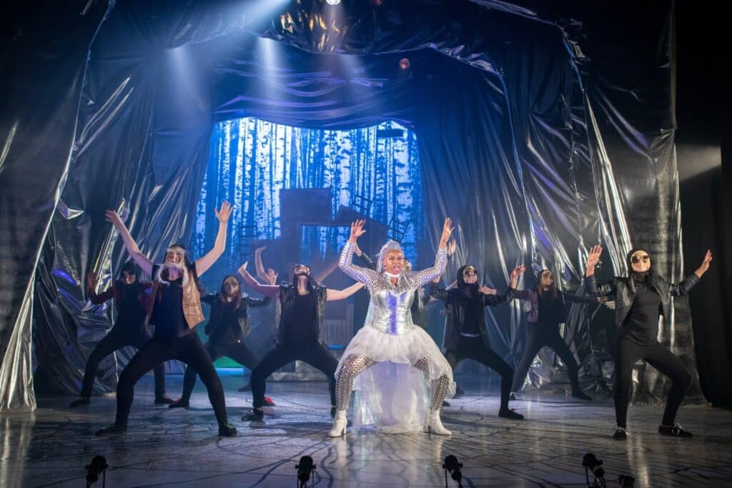 A performer in a silver, sparkling costume stands centre stage with arms raised, surrounded by dancers in black outfits and masks. The stage is draped in shiny fabric, and a backdrop shows a blue-toned forest scene, creating a dramatic, theatrical atmosphere.