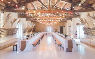 A rustic banqueting hall with high wooden beams, exposed stone walls, and colourful string lights. Two long tables covered in white cloths are lined with wooden benches, set for a formal meal, with plants and simple place settings as decorations.