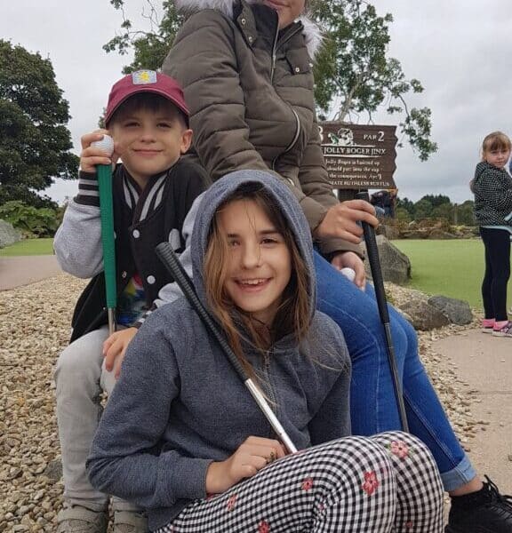 Three children in casual clothes sit outdoors on rocks, smiling and holding golf clubs. The background shows a golf course, a tree, a brown sign, and a cloudy sky. Another child stands in the distance on a green path.