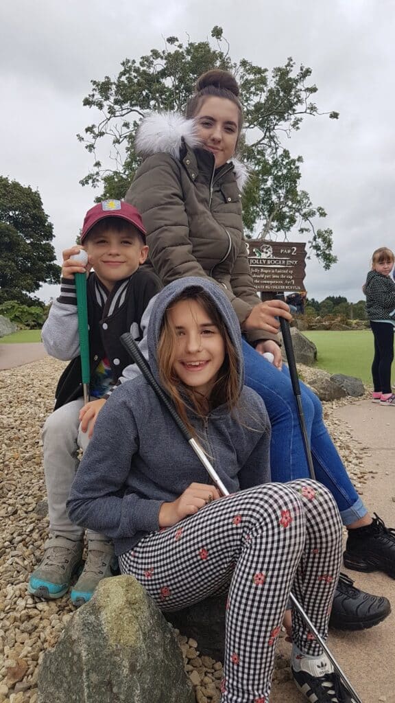 Three children sit on rocks at a mini-golf course, smiling at the camera. The girl at the front wears checked trousers and a grey hoodie. Behind her, a child in a cap and jacket holds a golf club, and a third child in a green coat sits higher up. A green lawn and trees are in the background.