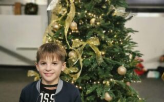 A young boy stands smiling in front of a decorated Christmas tree with gold ribbons and ornaments. He holds a golden ticket labelled "Return." The tree is topped with a large gold bow, and the indoor setting appears festive.