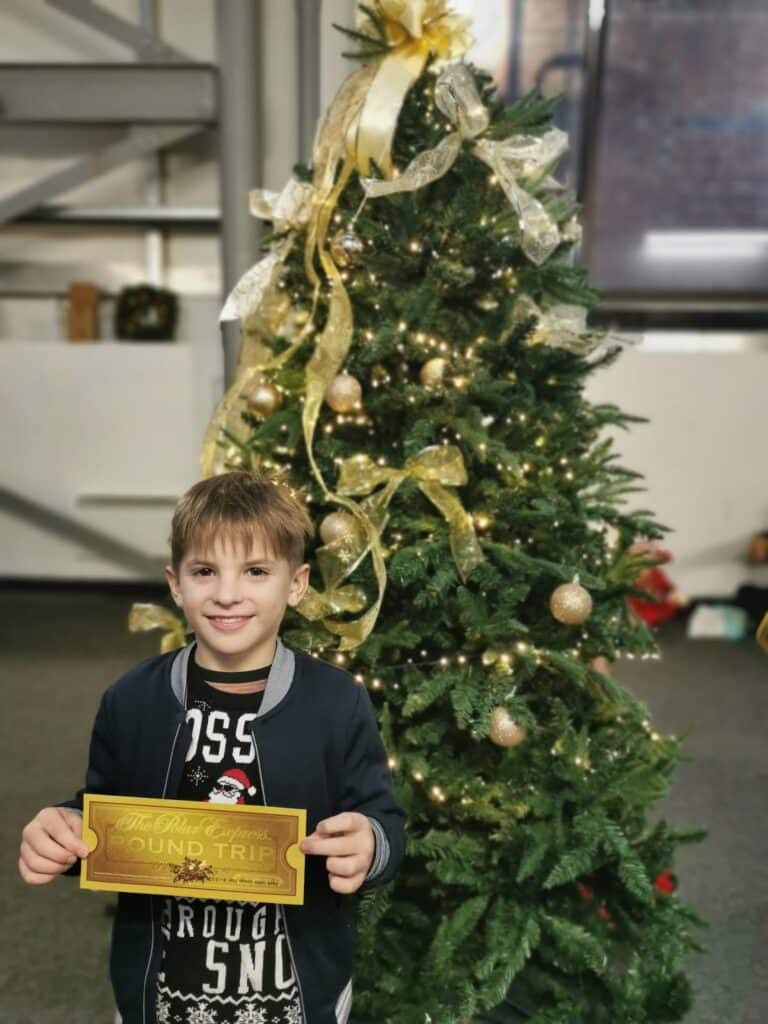 A smiling young boy stands in front of a decorated Christmas tree with gold ribbons and ornaments. He holds a gold "Return" ticket. The setting is indoors, with a window and shelves visible in the background.