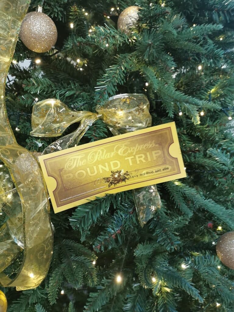 A golden “The Polar Express Return Ticket” hangs on a Christmas tree adorned with gold ribbon, gold baubles, and white fairy lights. The ticket features decorative designs, adding a festive touch to the holiday-themed tree.