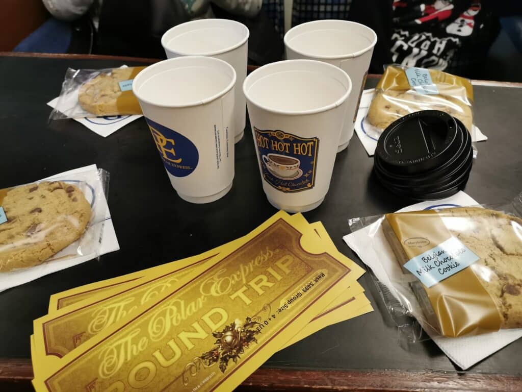 Three yellow "Polar Express Return Trip" tickets, three large chocolate chip biscuits in packaging, three white paper cups with hot beverage sleeves, and two black cup lids are on a dark tabletop. The scene suggests a festive or themed event.