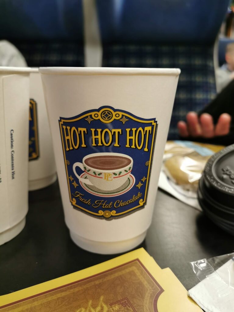 A white paper cup with a blue and gold badge that reads "HOT HOT HOT" above a steaming cup of hot chocolate. The text "Irish Hot Chocolate" appears below. The cup sits on a dark table, with snacks and other items blurred in the background.