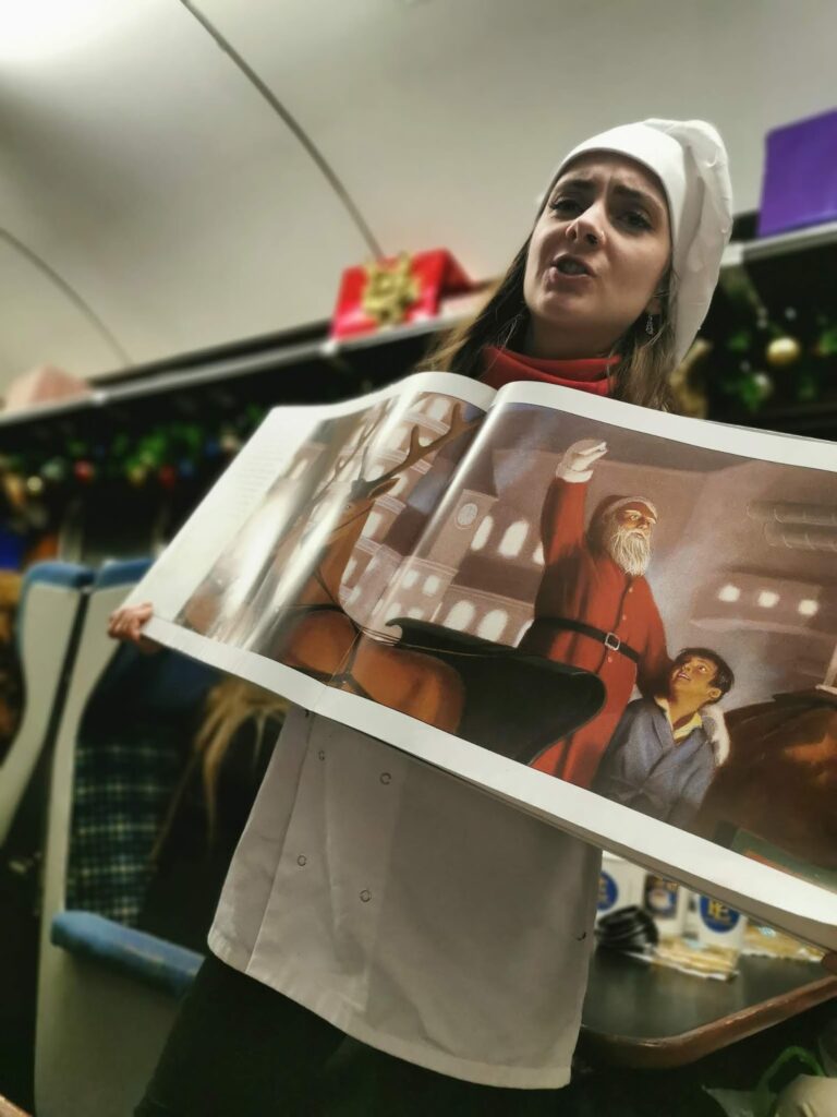 A woman dressed as a chef holds up an open picture book, showing an illustration of Father Christmas pointing while standing beside a boy. Festive decorations and presents are visible in the background, suggesting a holiday or Christmas-themed setting.