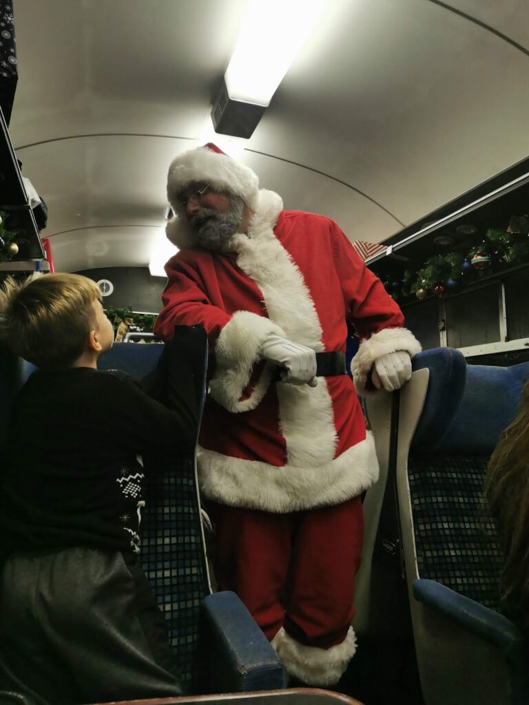 A man dressed as Father Christmas stands inside a train, leaning on a seat and smiling at a young boy. The boy looks up at Father Christmas with curiosity. The train is decorated with garlands and has blue checked seats. The lighting is warm and festive.