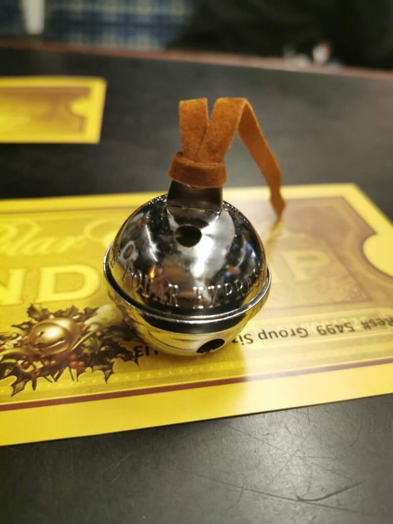A shiny silver bell with a brown suede strap sits on a yellow Polar Express-themed ticket. The bell reflects light, and the background shows a dark table surface and a blurred checked object.
