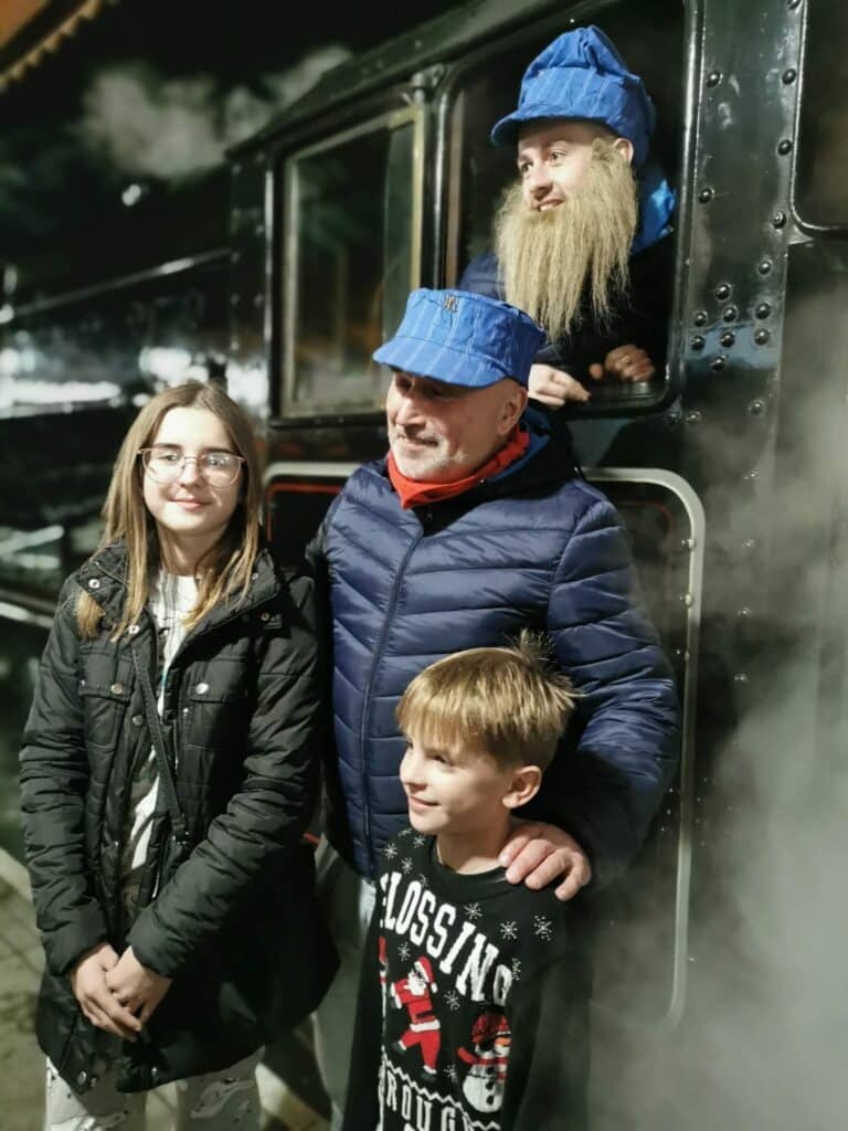 A man and two children stand smiling in front of a steam train. The man in blue is wearing a red scarf and blue cap. The girl wears glasses and a black jacket, whilst the boy wears a festive jumper. A person with a fake beard and blue hat leans from the train window above.