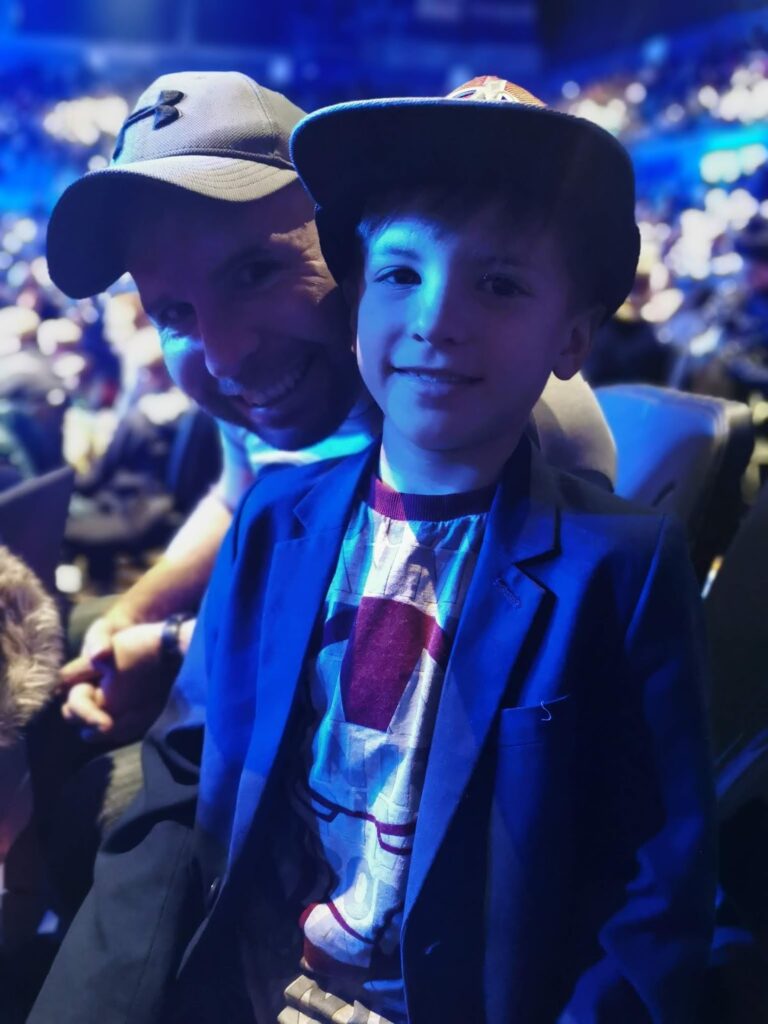 A man in a cap and a young boy in a blue blazer and cap smile at the camera in a crowded, brightly lit indoor arena, with blue lighting and blurry spectators in the background.
