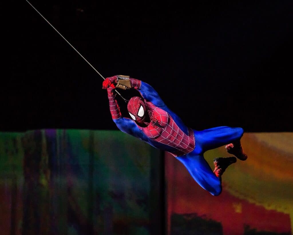 A person dressed as Spider-Man in a red and blue suit with web patterns swings through the air on a web, against a colourful, blurred background. The character’s body is angled horizontally, legs bent, and both hands gripping the web above.