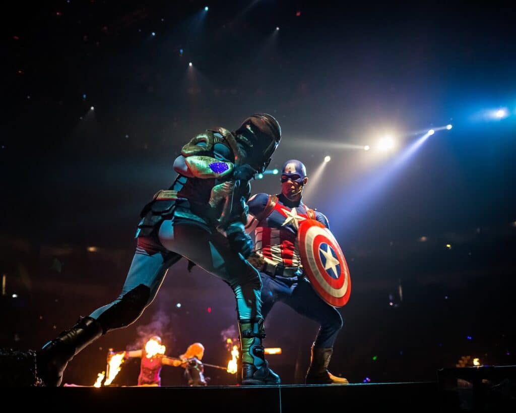 Two costumed performers dressed as Marvel superheroes Captain America and Star-Lord pose in dramatic, action-ready stances on a stage with spotlights and fire effects in the background, creating an epic, high-energy atmosphere.