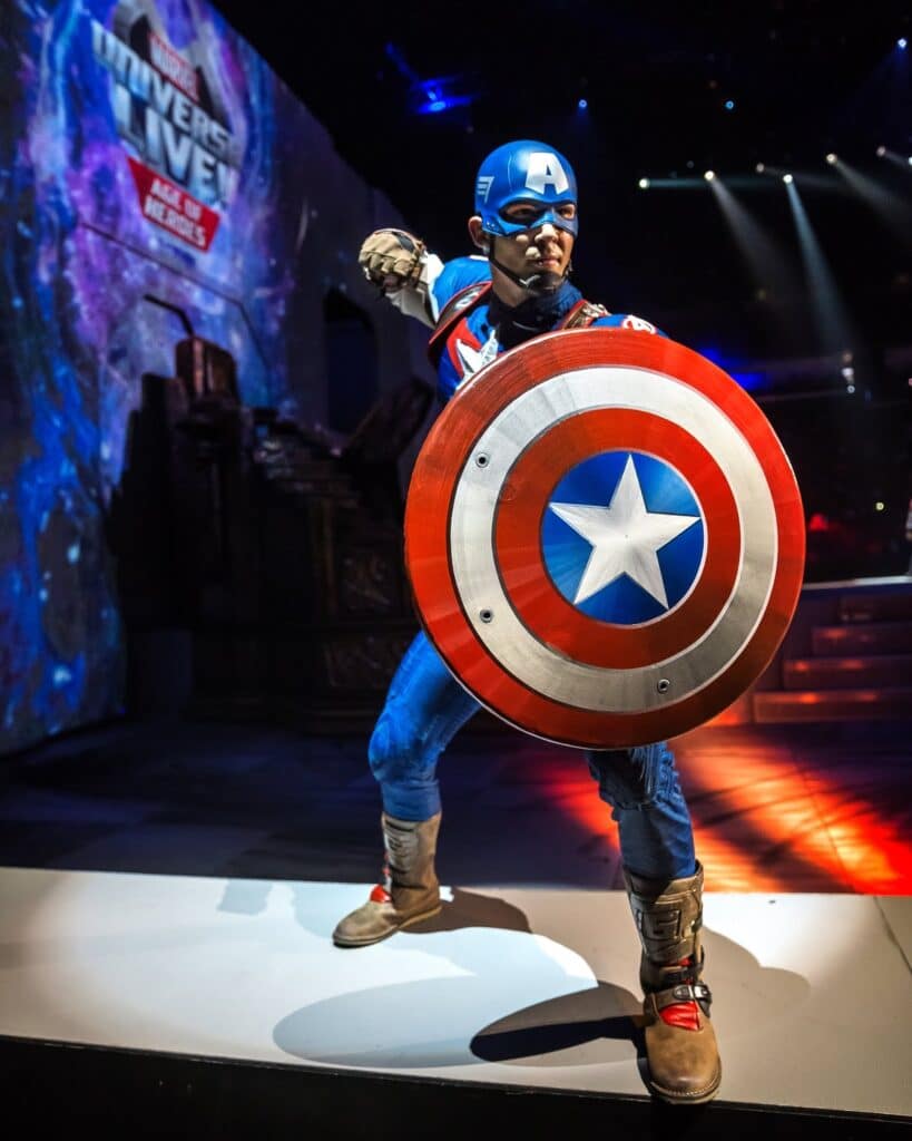 A person dressed as Captain America stands on a stage, holding a red, white, and blue shield. The background is dark with dramatic lighting and a large "Marvel Universe Live" sign is visible on the left side. The pose is dynamic and heroic.