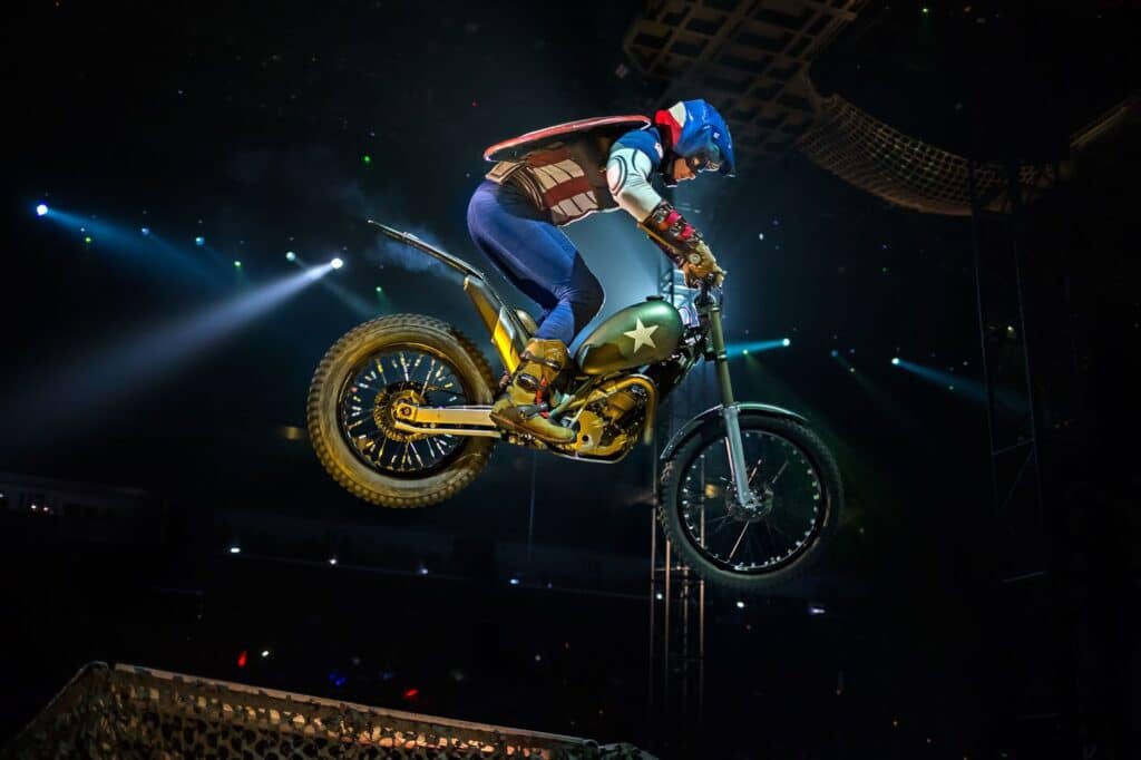 A motorcyclist in a Captain America costume jumps a dirt bike off a ramp indoors. The rider wears a blue helmet, red gloves, and a star-spangled jacket. The bike has knobbly tyres and a green body with a white star. Spotlights and a stage structure are visible in the background.