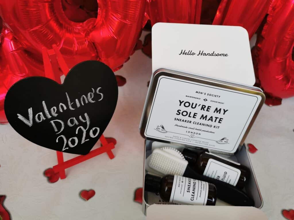 A trainer cleaning kit in an open box labelled “You’re My Sole Mate” sits beside a heart-shaped chalkboard on a mini easel that reads “Valentine’s Day 2020.” Red heart balloons are in the background, creating a festive atmosphere.