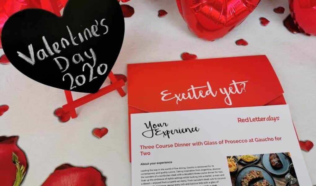 A red and white card advertising a three-course dinner with prosecco is on a table decorated with red rose petals and heart balloons. A black heart-shaped chalkboard on a mini easel reads “Valentine’s Day 2020” in white script.