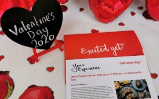 A red and white card advertising a three-course dinner with prosecco is on a table decorated with red rose petals and heart balloons. A black heart-shaped chalkboard on a mini easel reads “Valentine’s Day 2020” in white script.