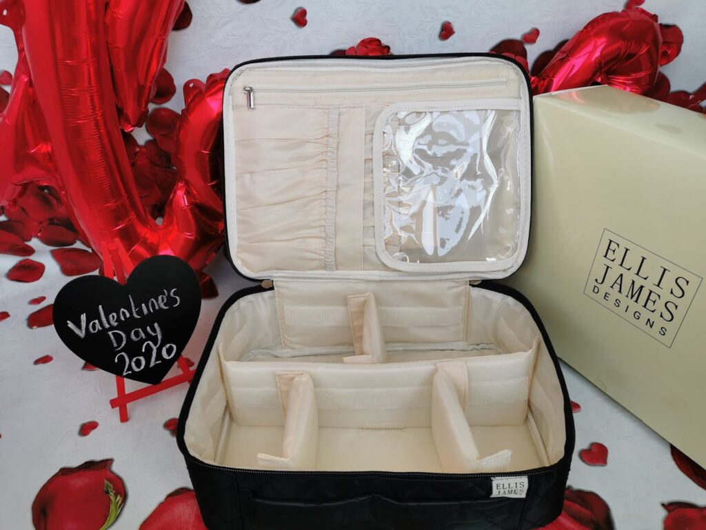 An open black quilted organiser with beige interior dividers and clear zippered pockets sits on a rose petal-patterned surface, next to an Ellis James Designs box. A black heart sign reads "Valentine's Day 2020" with red balloon decorations in the background.