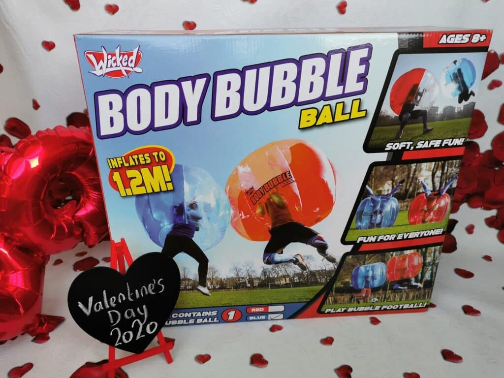 A boxed “Wicked Body Bubble Ball” toy is displayed, featuring images of children playing inside large inflatable blue and red balls. The box says it inflates to 1.2m, is for ages 8+, and highlights features. A heart sign reads “Valentine’s Day 2020.” Red decorations surround it.