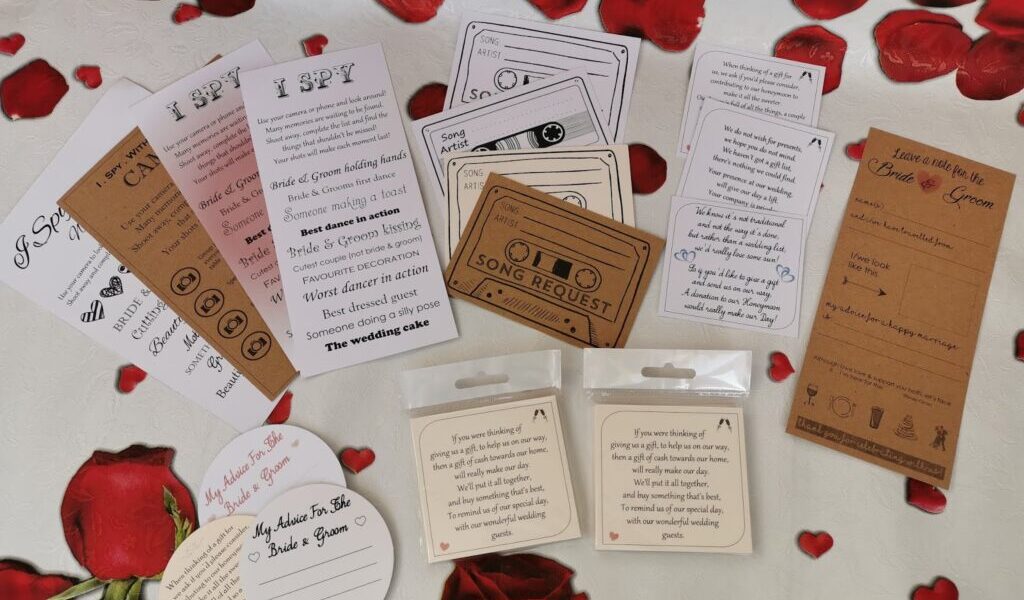 A collection of wedding-themed cards—“I Spy”, song request, advice cards, and others—are spread out on a white surface with scattered red rose petals and printed roses. The cards are in different shapes, sizes, and colours, including white, brown, and blush pink.