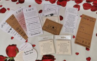 A collection of wedding-themed cards—“I Spy”, song request, advice cards, and others—are spread out on a white surface with scattered red rose petals and printed roses. The cards are in different shapes, sizes, and colours, including white, brown, and blush pink.