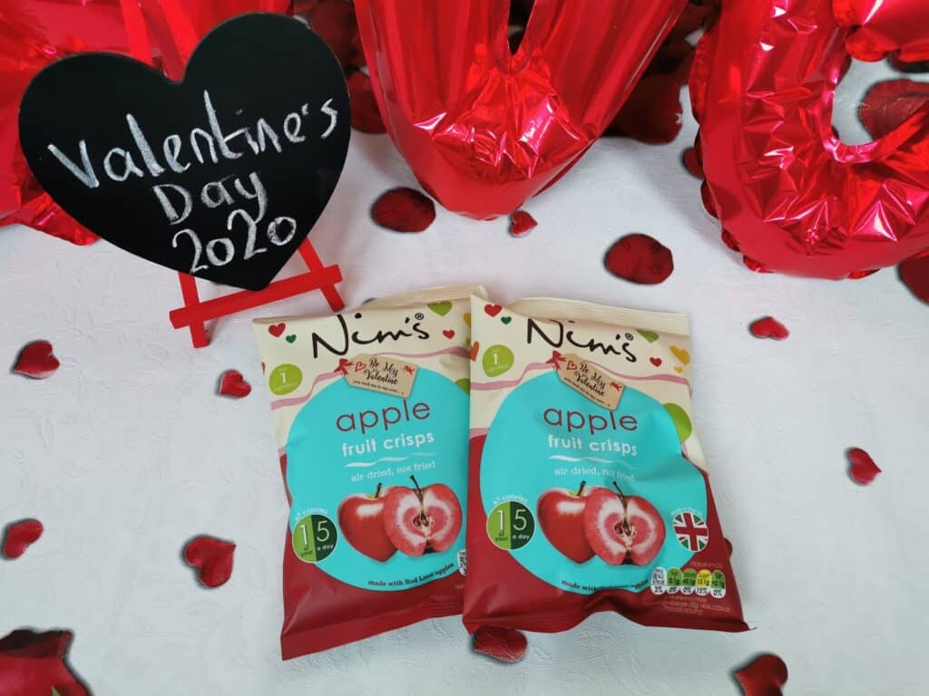 Two packets of Nim’s apple fruit crisps sit on a white surface scattered with red heart confetti. Behind them are red balloon letters spelling "LOVE" and a blackboard heart sign with "Valentine's Day 2020" written in white.
