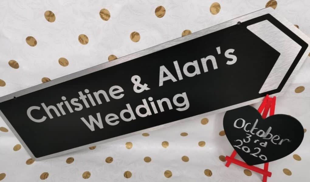 A black sign with white text reads “Christine & Alan’s Wedding” and points right. Beside it, a small black heart on a red easel displays “3rd October 2020.” The background is white with gold polka dots.