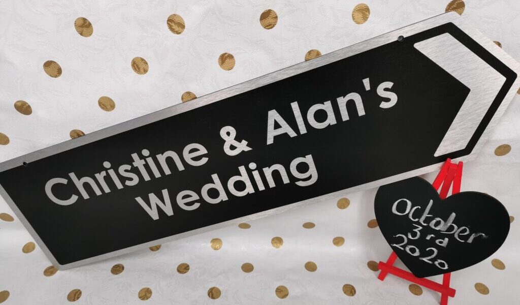 A black sign with white text reads “Christine & Alan’s Wedding” and points right. Beside it, a small black heart on a red easel displays “3rd October 2020.” The background is white with gold polka dots.