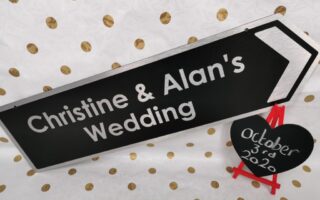 A black sign with white text reads “Christine & Alan’s Wedding” and points right. Beside it, a small black heart on a red easel displays “3rd October 2020.” The background is white with gold polka dots.