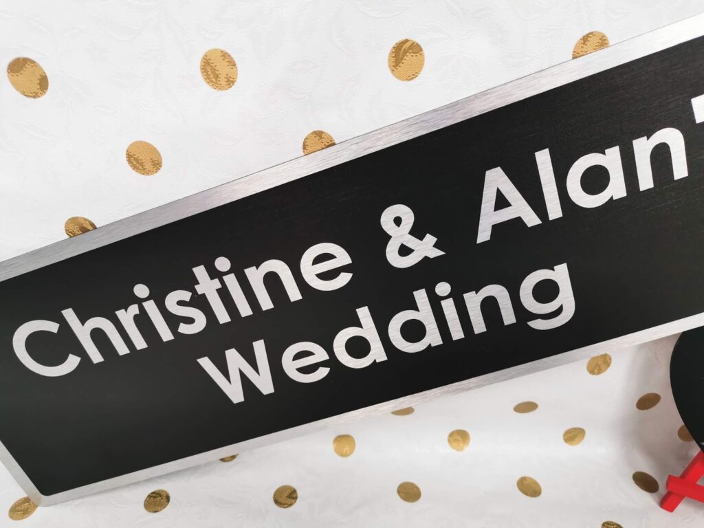A rectangular black sign with silver edges and bold white text reads “Christine & Alan Wedding.” The background is white with large gold spots. Only part of the sign is visible, and the lower right corner shows a glimpse of a red object.