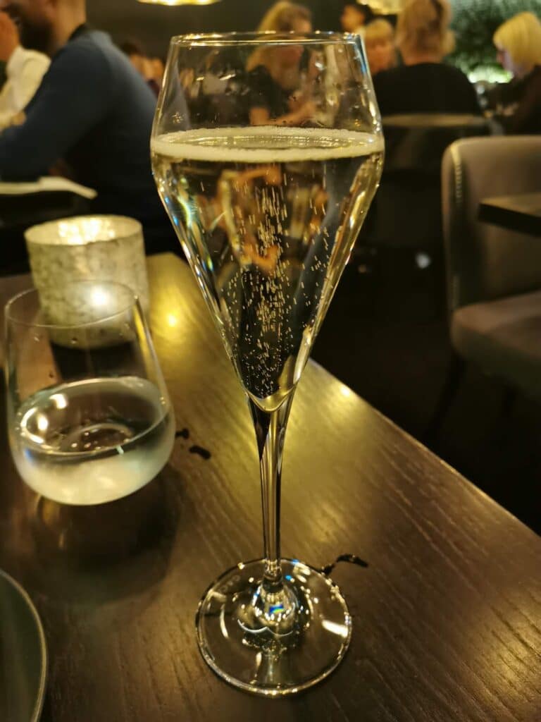 A tall champagne flute filled with sparkling wine sits on a dark wooden table. Bubbles rise in the glass. Nearby are a lit candle in a stone holder and a glass of water. The blurred background shows people dining in a warmly lit restaurant.