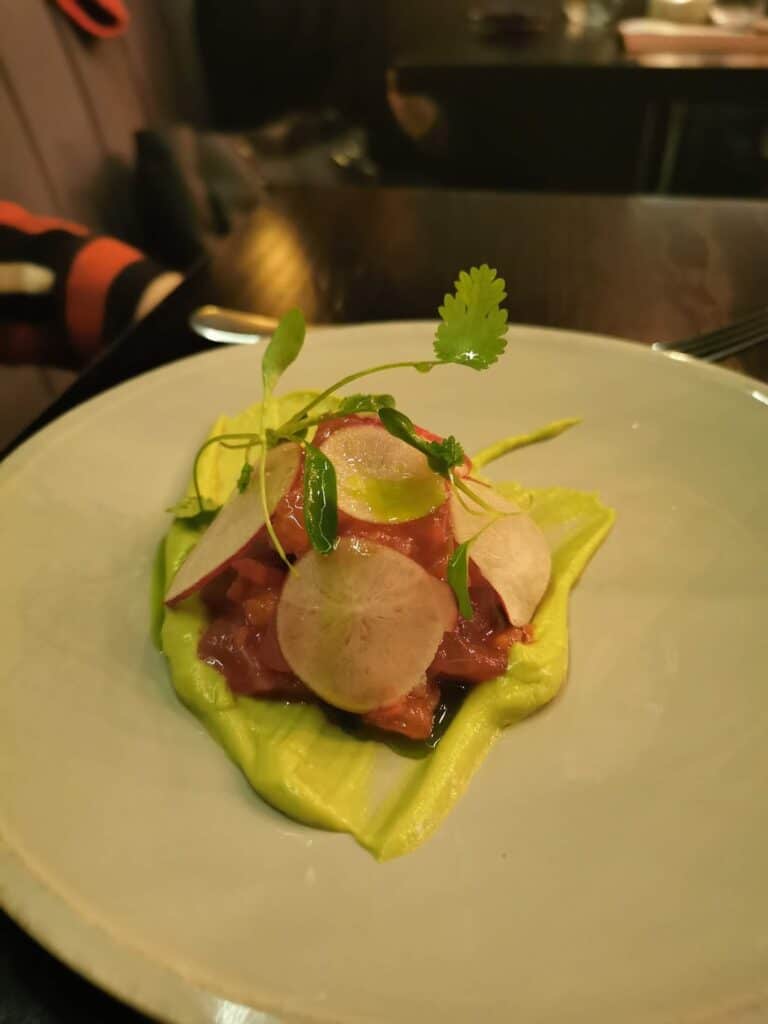 A white plate holds a mound of diced tuna tartare on a bed of creamy green avocado, topped with thinly sliced radishes and delicate microgreens, presented on a dark restaurant table with dim, warm lighting.
