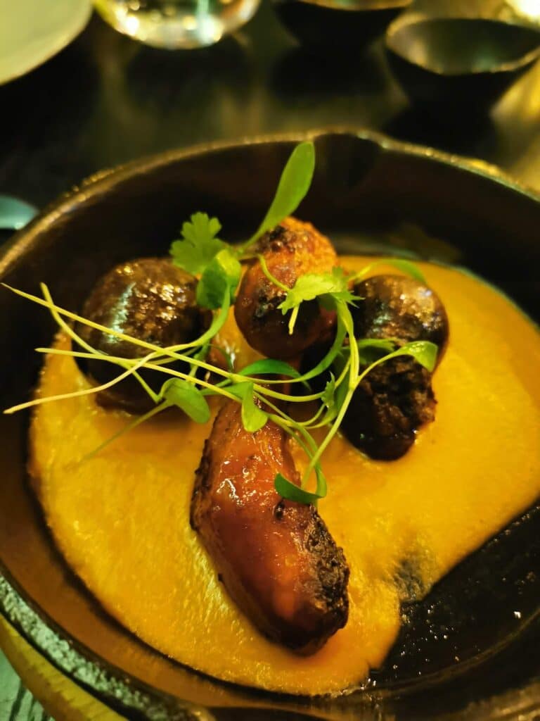 A black bowl contains three roasted mushrooms on a bed of creamy orange sauce, garnished with fresh green microgreens and coriander. The background shows blurred bowls and glasses on a dark, reflective table.