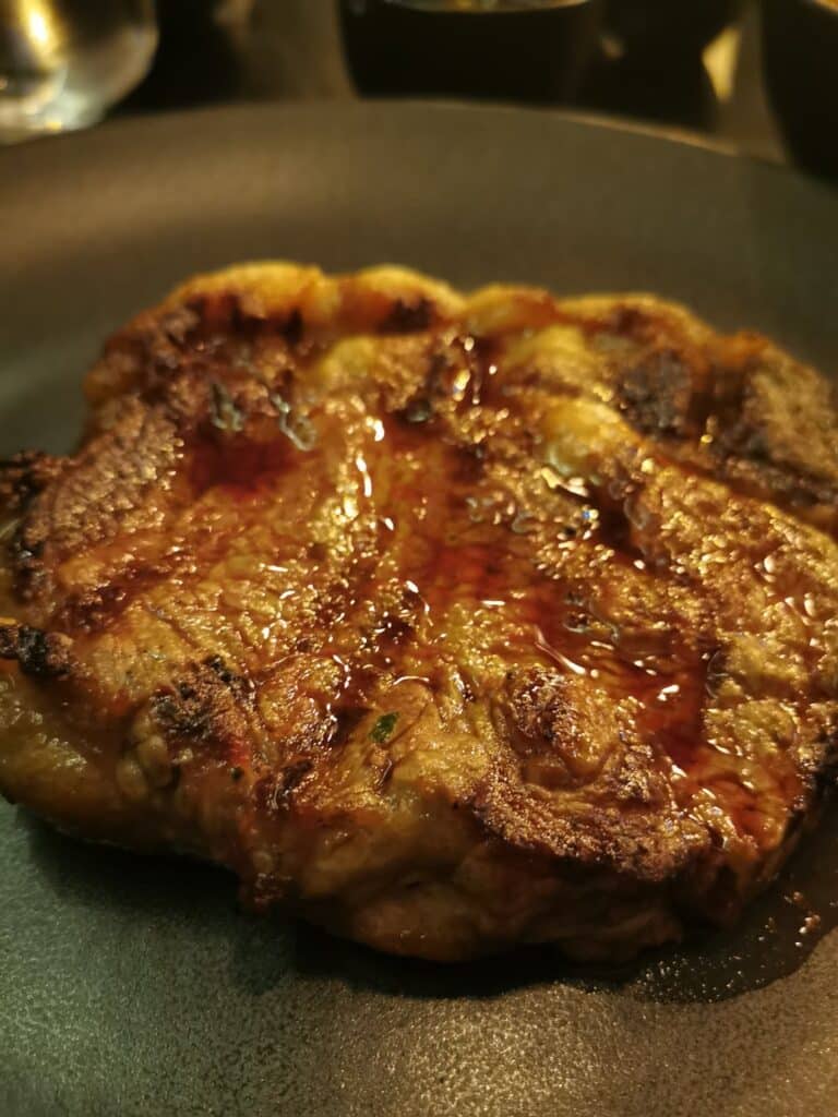 A close-up of a cooked steak on a dark plate. The steak has a browned, slightly charred surface with visible grill marks and a glistening layer of juices, indicating it is freshly cooked and moist. The background is softly blurred.