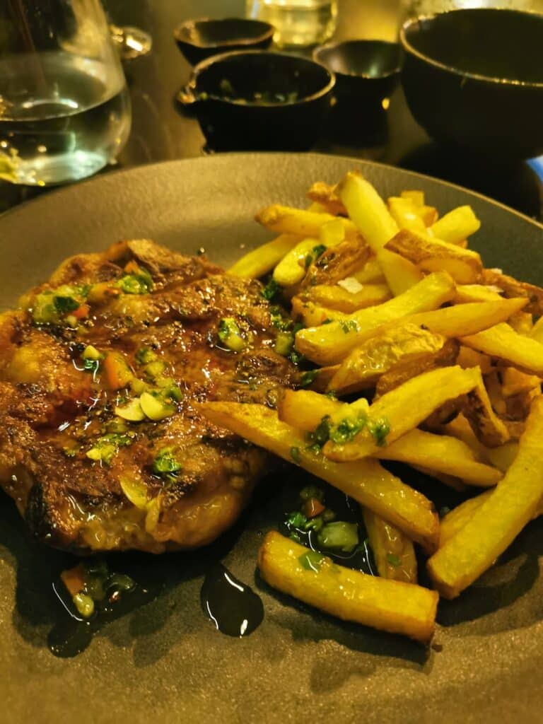 A dark plate with grilled steak topped with herbs and sauce is served beside golden, crispy chips. Small bowls with condiments and a glass of water are in the background on a reflective table.