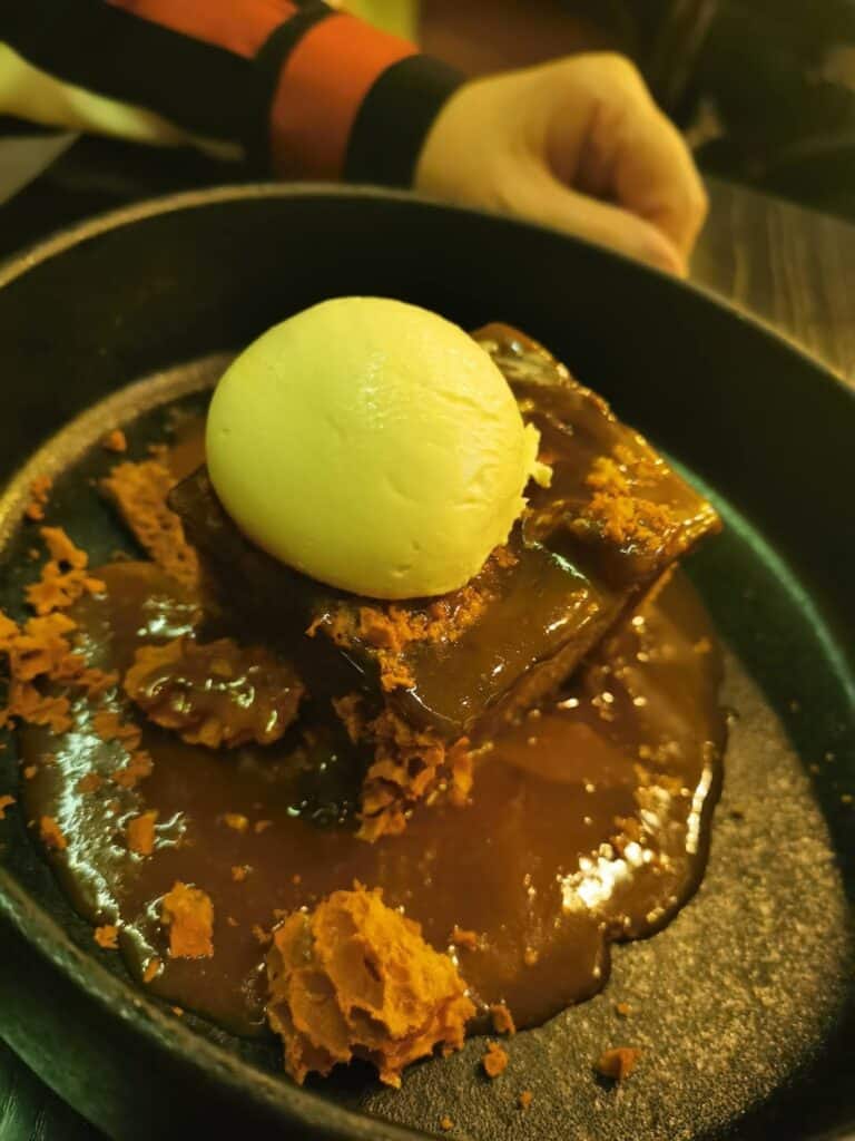 A close-up of a dessert in a black dish features a square slice of sticky toffee pudding topped with a scoop of vanilla ice cream and surrounded by crumbled honeycomb and rich toffee sauce. A hand rests in the background, partially visible above the dessert.