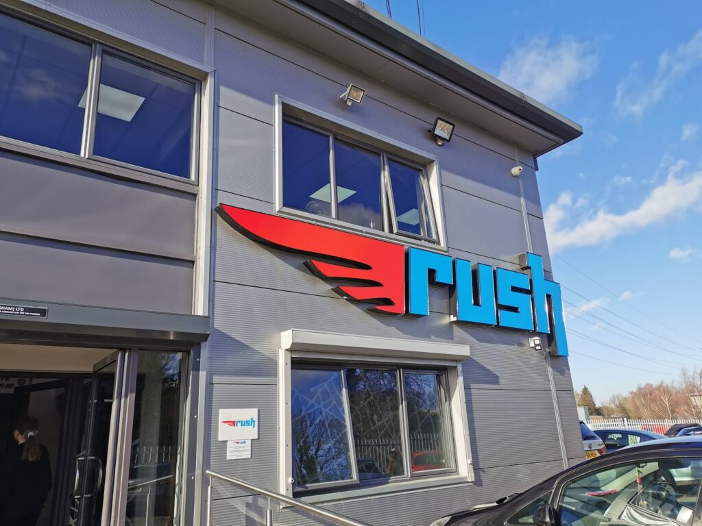 A modern building exterior with large windows and a sign that reads "Rush" in bold blue and red letters, with a red wing design extending from the "R." The sky is blue with some clouds, and cars are parked outside.