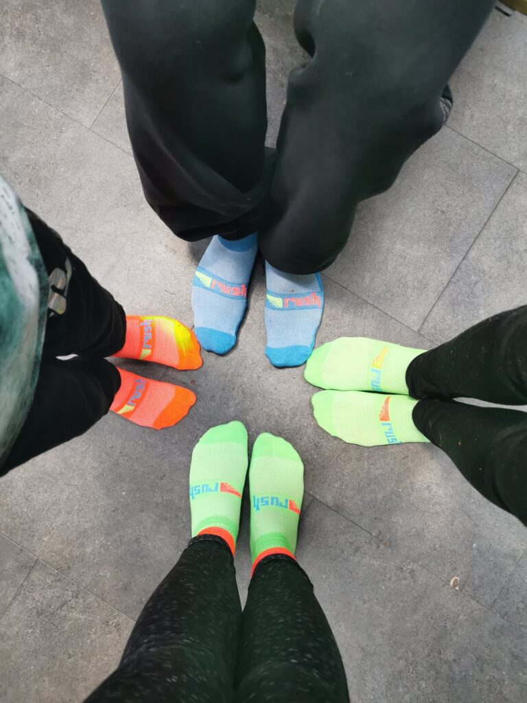 Four people stand in a circle on a grey tiled floor, each wearing colourful socks. The socks are bright orange, blue, green, or yellow, with the same brand name printed near the toes. The people’s legs are dressed in dark trousers.