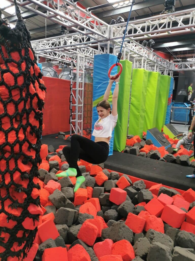 A young woman in a white shirt and black leggings hangs from a red ring over a pit filled with orange and grey foam blocks in an indoor obstacle course, surrounded by nets, padded pillars, and metal frames. She looks focused and mid-swing.
