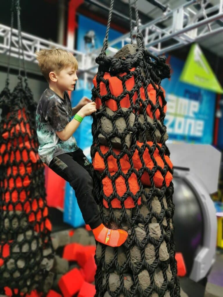 A young boy wearing a black and grey shirt, black trousers, and orange socks climbs a large rope net structure with red and black sections at an indoor play centre. Red foam blocks are scattered on the floor below him. The background shows blurred play equipment.